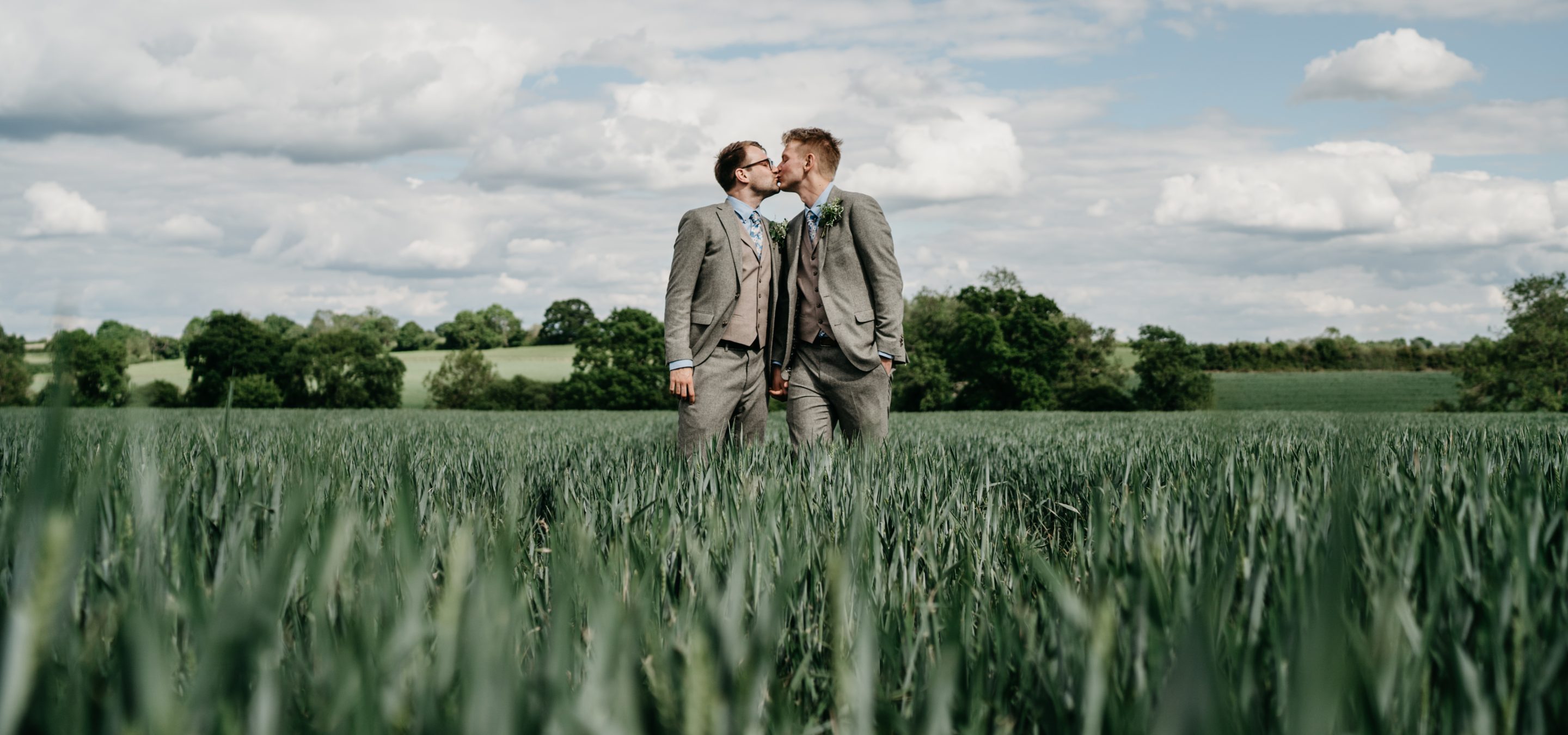 Two grooms sharing a kiss in a lush green field at Huntsmill Farm sustainable wedding venue